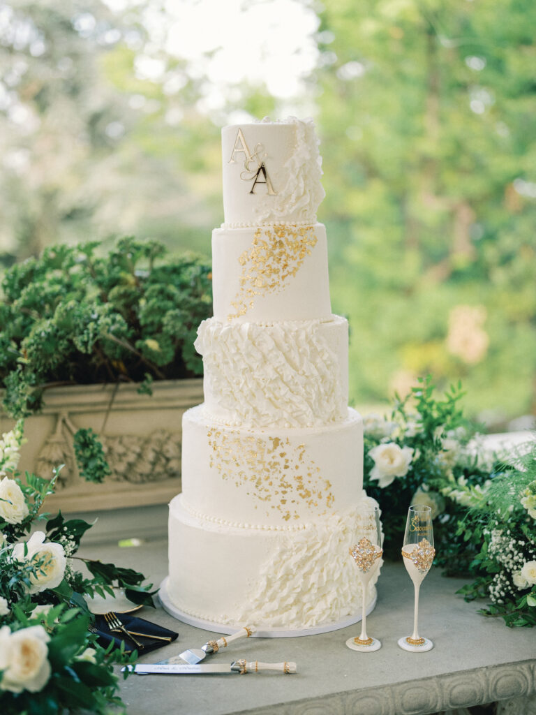 Five-tier white wedding cake with gold leaf texture, elegant monogram, and ruffled frosting details.