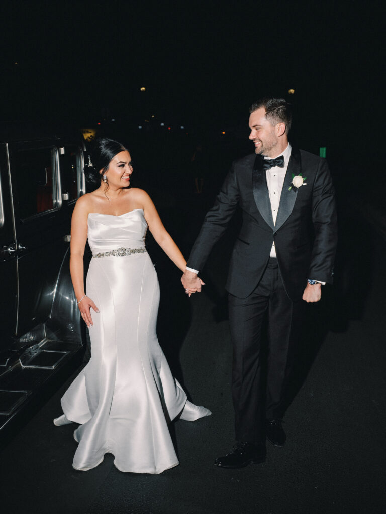 Bride and groom holding hands and laughing during a night wedding exit next to a vintage getaway car