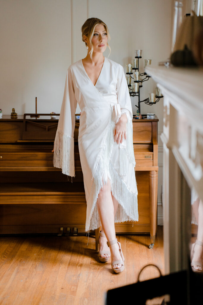 Bride in an elegant silk wrap robe with fringe details leaning against a vintage wooden piano.