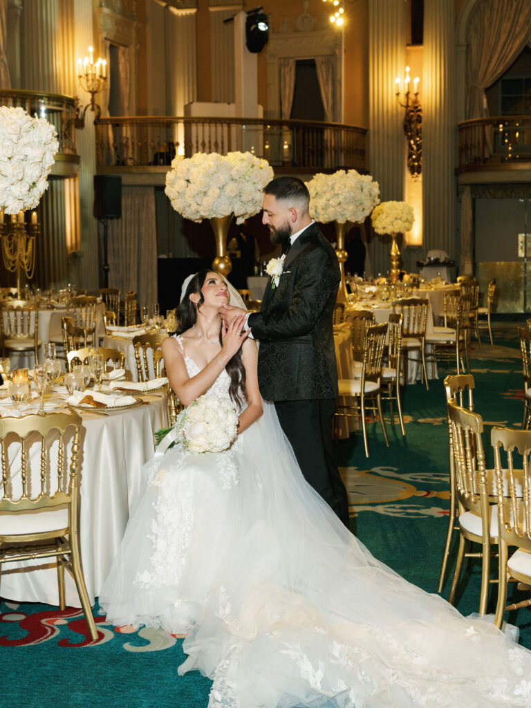 A newlywed couple shares an intimate moment in an opulent Los Angeles ballroom with gold chiavari chairs and white floral centerpieces.