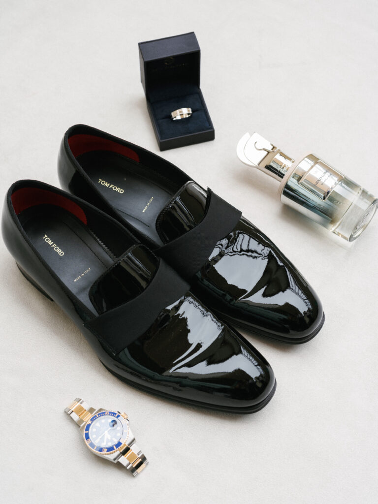 Tom Ford black patent leather loafers styled on a neutral surface with a gold Rolex watch, luxury perfume bottle, and a wedding ring box.