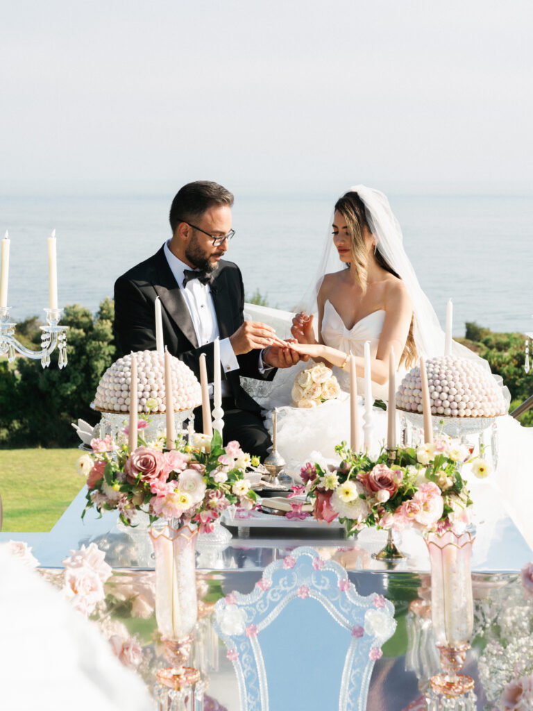 Groom placing a ring on the bride's finger during a traditional Persian Sofreh Aghd oceanfront ceremony.