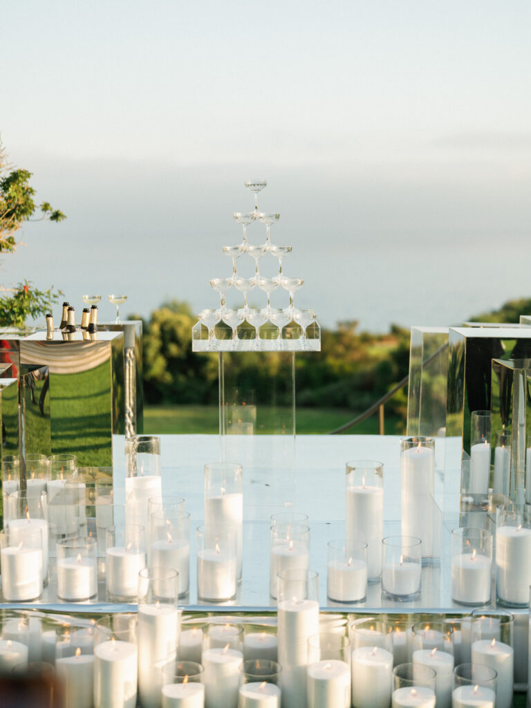 Champagne glass tower on a transparent stand surrounded by lit white candles at an outdoor wedding reception