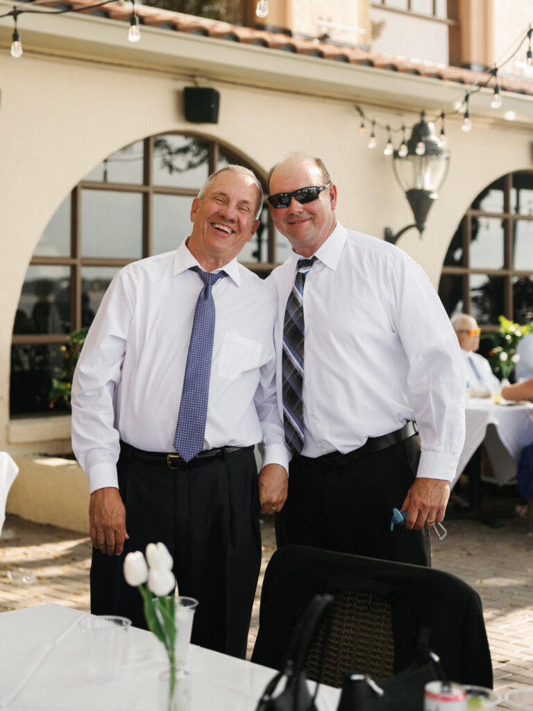 Two smiling male wedding guests in white dress shirts and ties posing together at an outdoor reception table decorated with white tulips and bistro string lights.