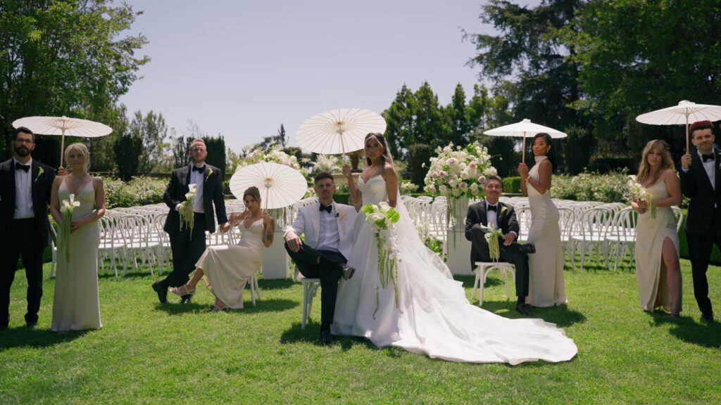A bride, groom, and wedding party in elegant formal attire and holding parasols pose for a group photo during their outdoor multicultural and traditional celebration.