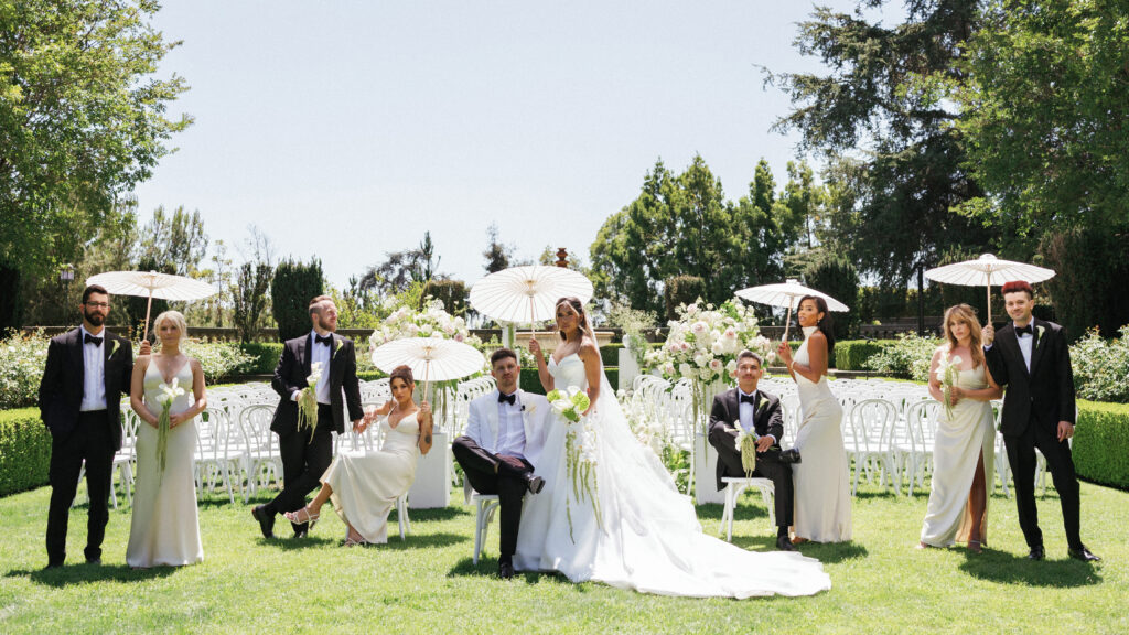 Editorial wedding party portrait featuring bridesmaids and groomsmen holding white parasols at an outdoor ceremony.