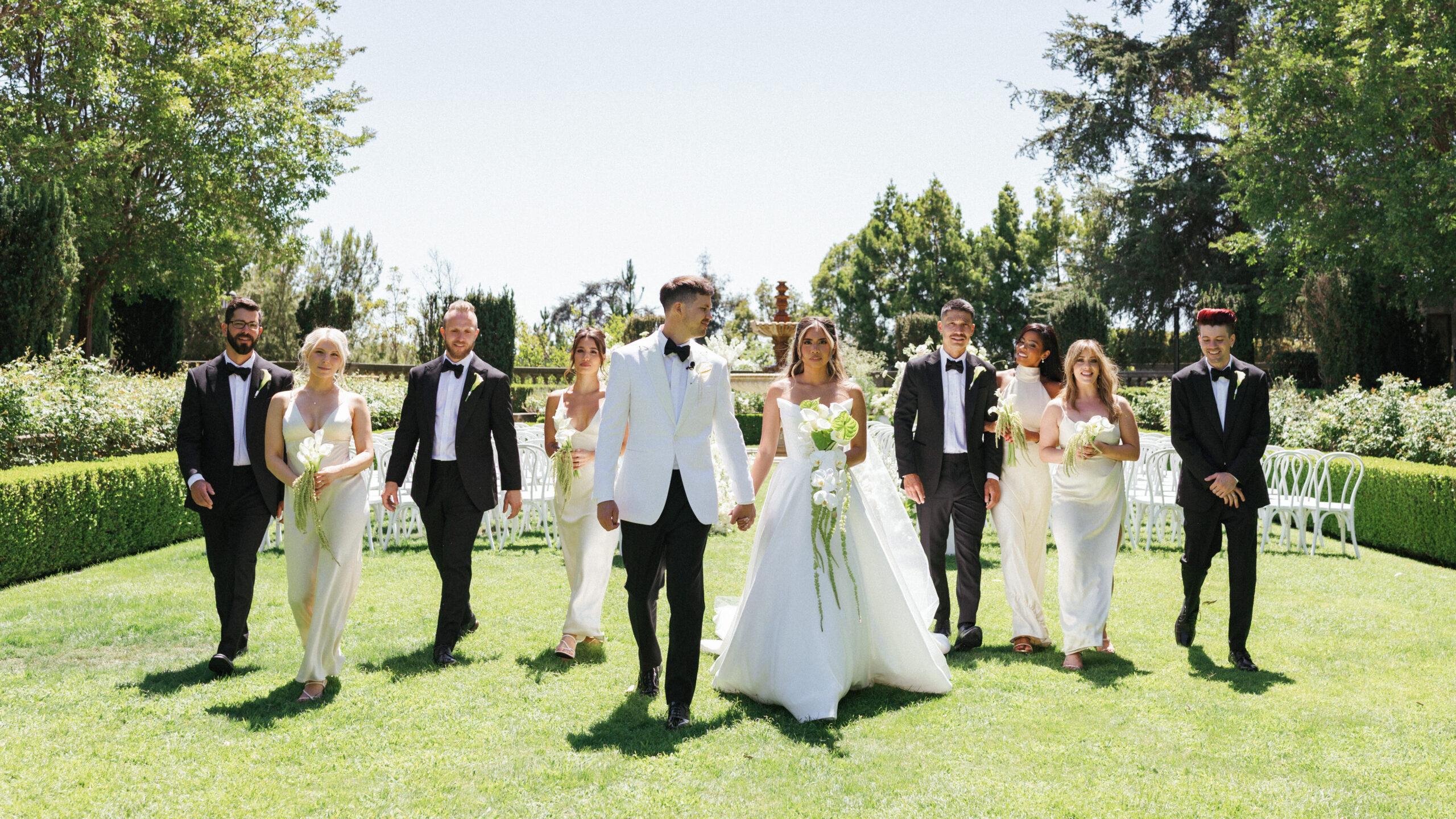 High-end outdoor family and bridal party wedding photo, captured in motion on a sun-drenched lawn of a California private estate with sophisticated black-tie and neutral attire.