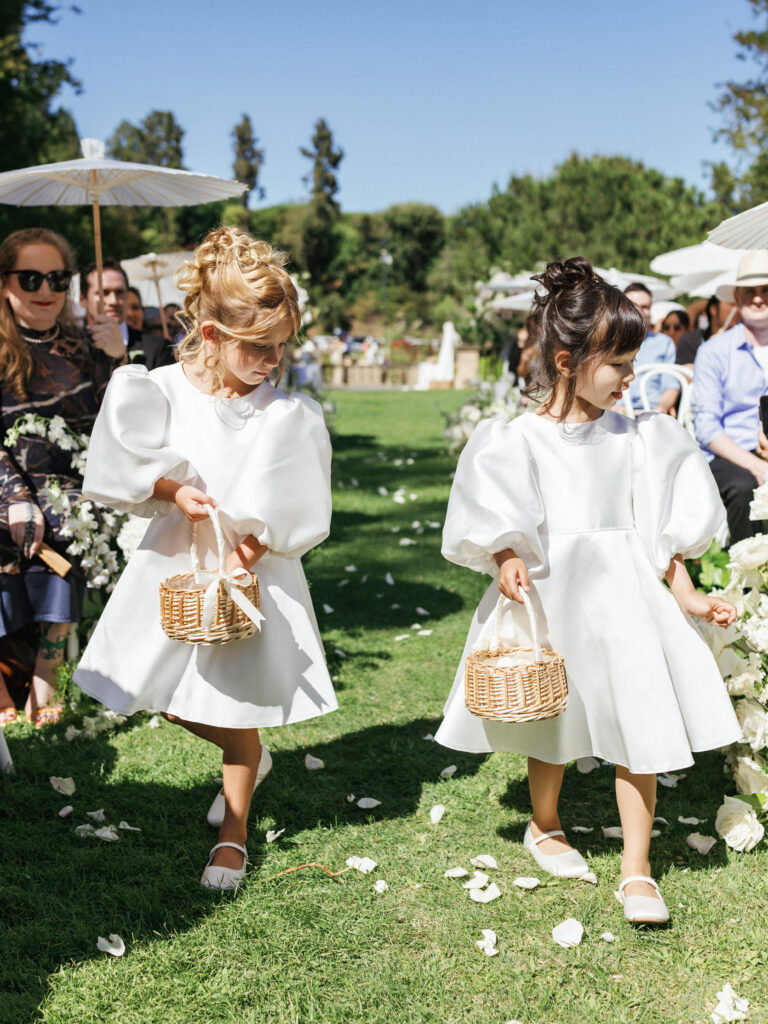 Flower girls in white dresses holding small baskets at a garden wedding ceremony.