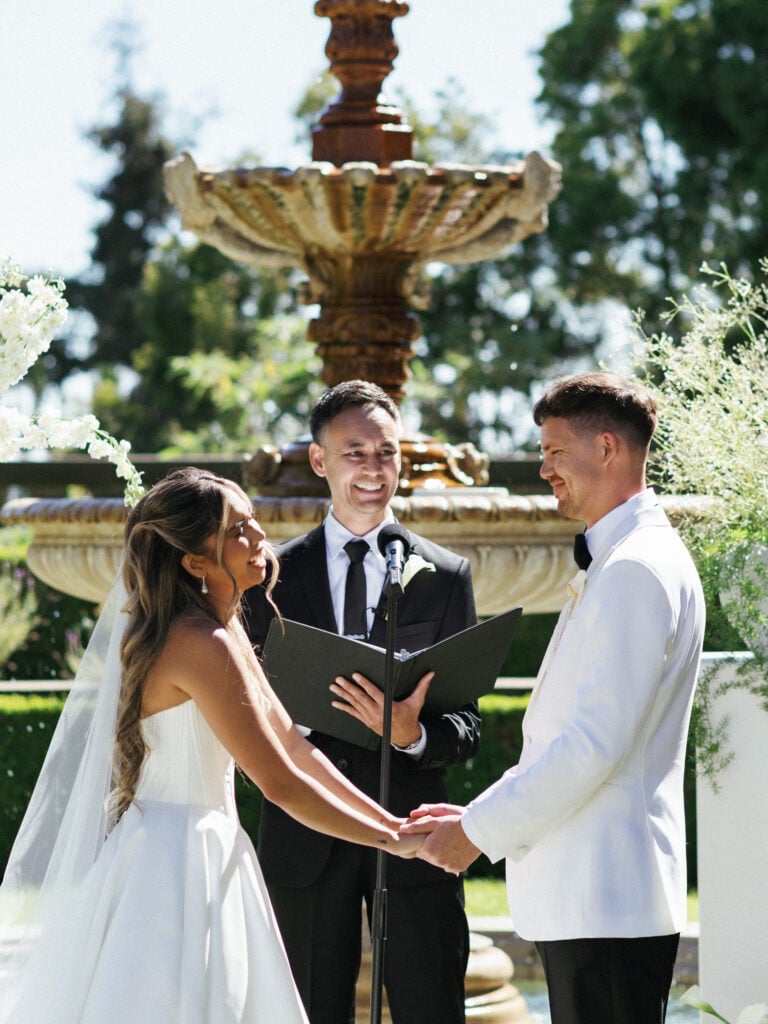 A bride and a groom in a white tuxedo jacket holding hands and facing each other during an outdoor wedding ceremony next to a large stone fountain.