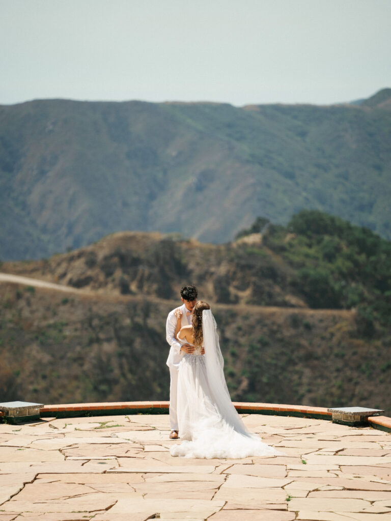 Drone wedding cinematography perspective showing a bride and groom embracing on a mountain cliffside stone terrace overlooking scenic canyon views.