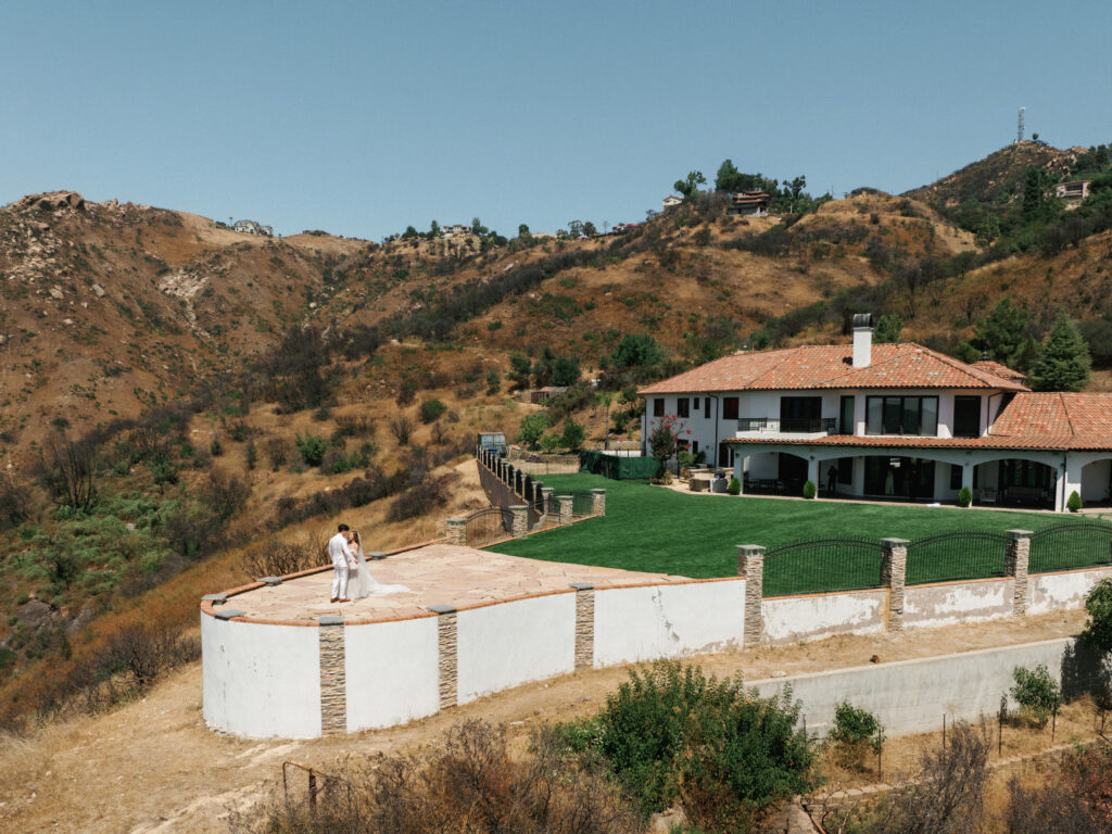Drone wedding cinematography wide shot of a luxury private Malibu villa and green lawn with newlyweds posing on an outdoor stone patio.