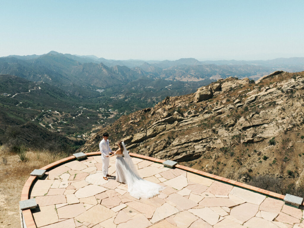 Aerial view drone wedding photography at a luxury Malibu mountain canyon venue with newlyweds holding hands on a cliffside stone overlook.