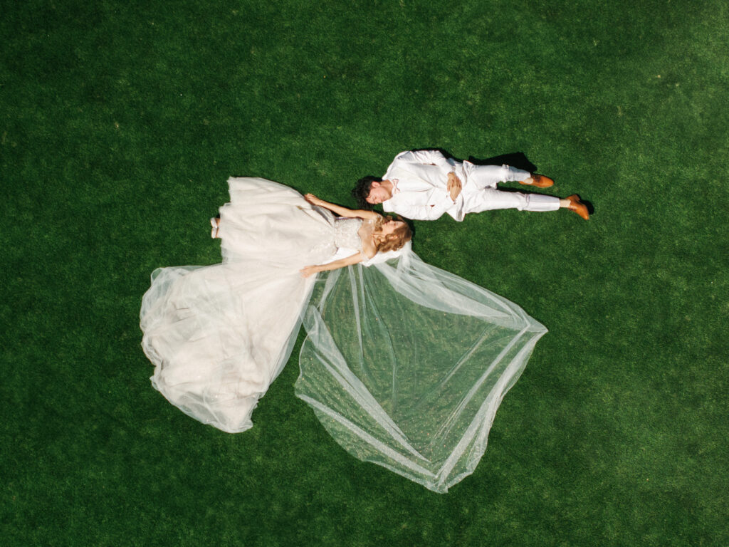 Top down drone wedding portrait of a bride in an A-line tulle gown with an extended long veil and groom in a white suit lying on a luxury estate green lawn.