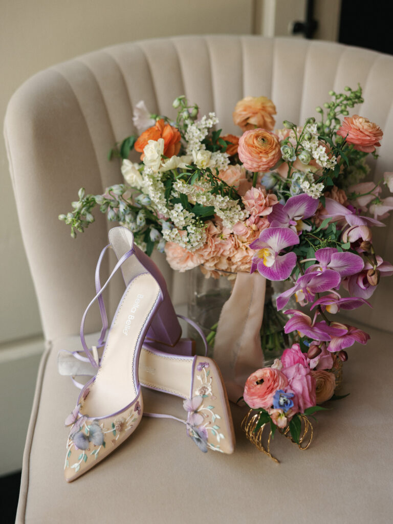 Lavender embroidered floral bridal heels leaning against a luxury wedding bouquet with purple orchids and peach ranunculus on a beige velvet armchair.
