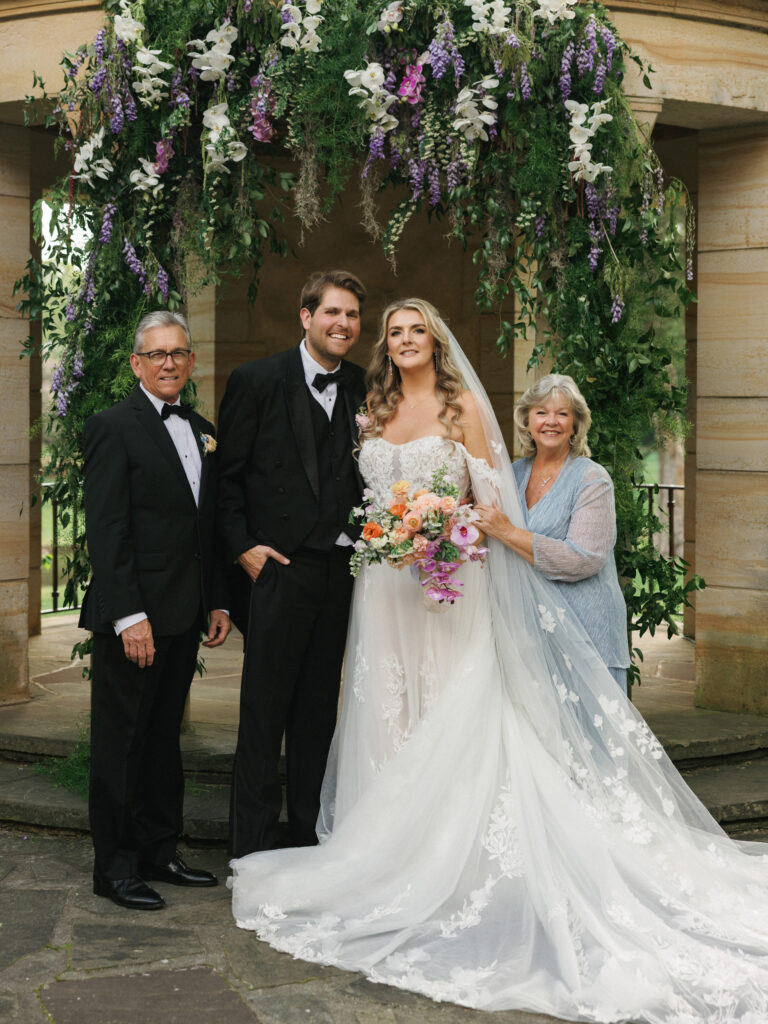 Editorial family wedding portrait under a lush stone gazebo installation filled with cascading lavender and white florals, showcasing a romantic bride and groom with family.