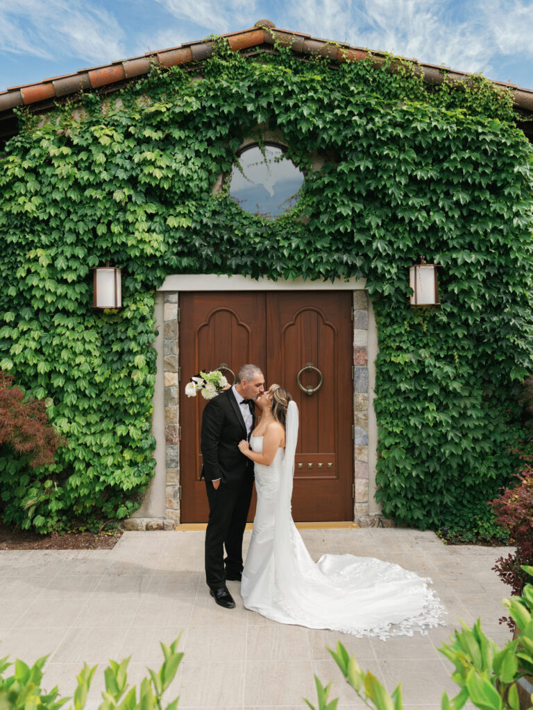 Newlyweds kissing in front of large wooden doors on an ivy-covered stone estate facade, showcasing the bride's long lace-trimmed train.