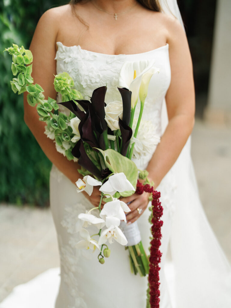 Close-up of a modern bridal bouquet featuring dark calla lilies, white orchids, and cascading amaranth, held by a bride in a strapless textured wedding gown.