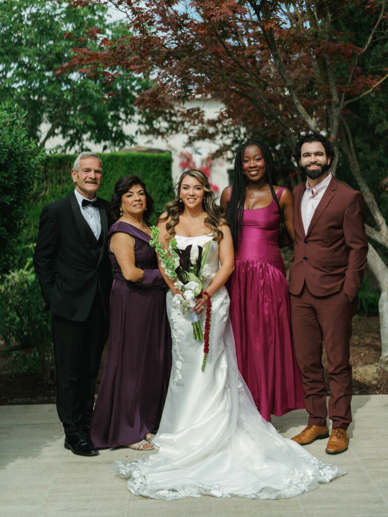 Bride in a satin wedding dress holding a dark calla lily bouquet, posing with her parents in a plum dress and tuxedo, and guests in a burgundy suit under a Japanese maple tree.