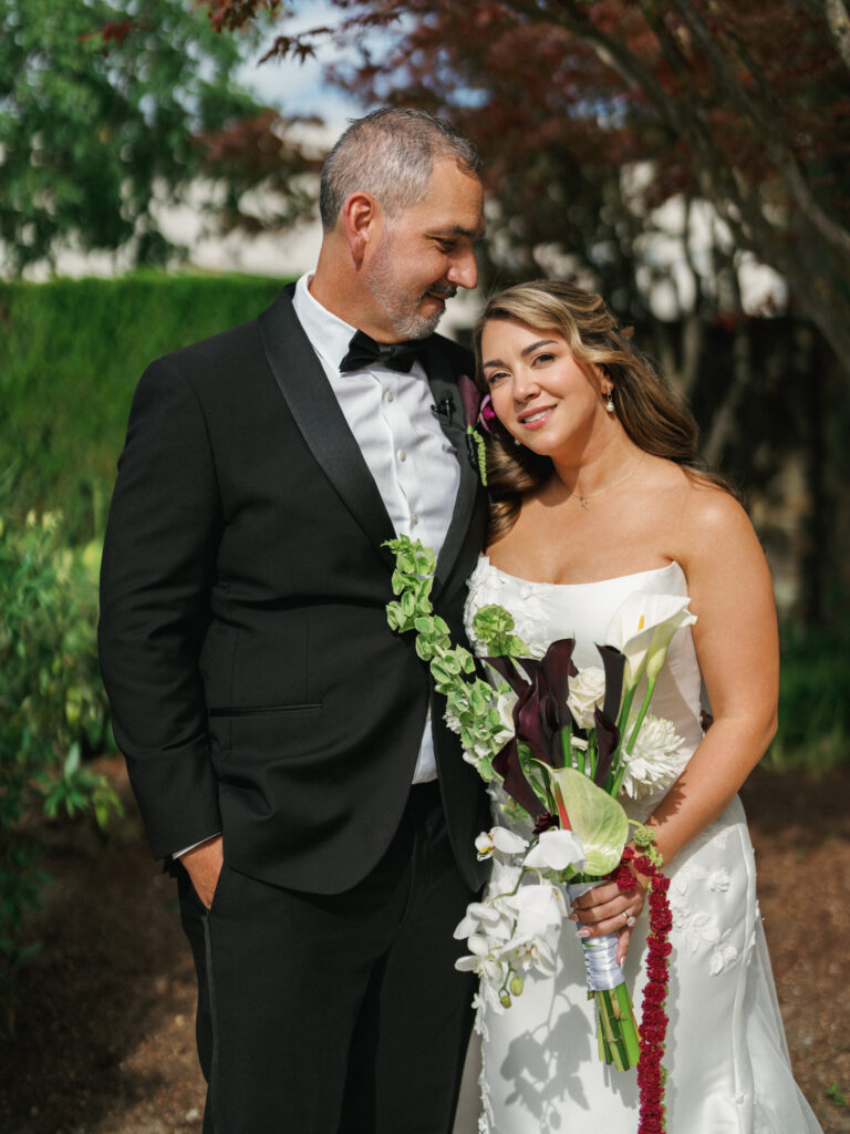 Outdoor wedding couple portrait, featuring a bride in a strapless dress with romantic downdo hairstyle and a groom in formal black-tie attire.