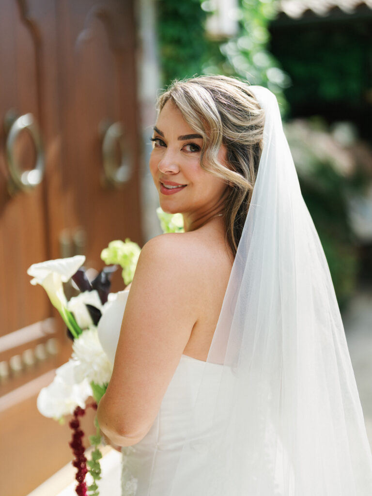 Bride with a half-up wedding hairstyle and a long tulle veil looking over her shoulder while holding a white calla lily bouquet by wooden estate doors.