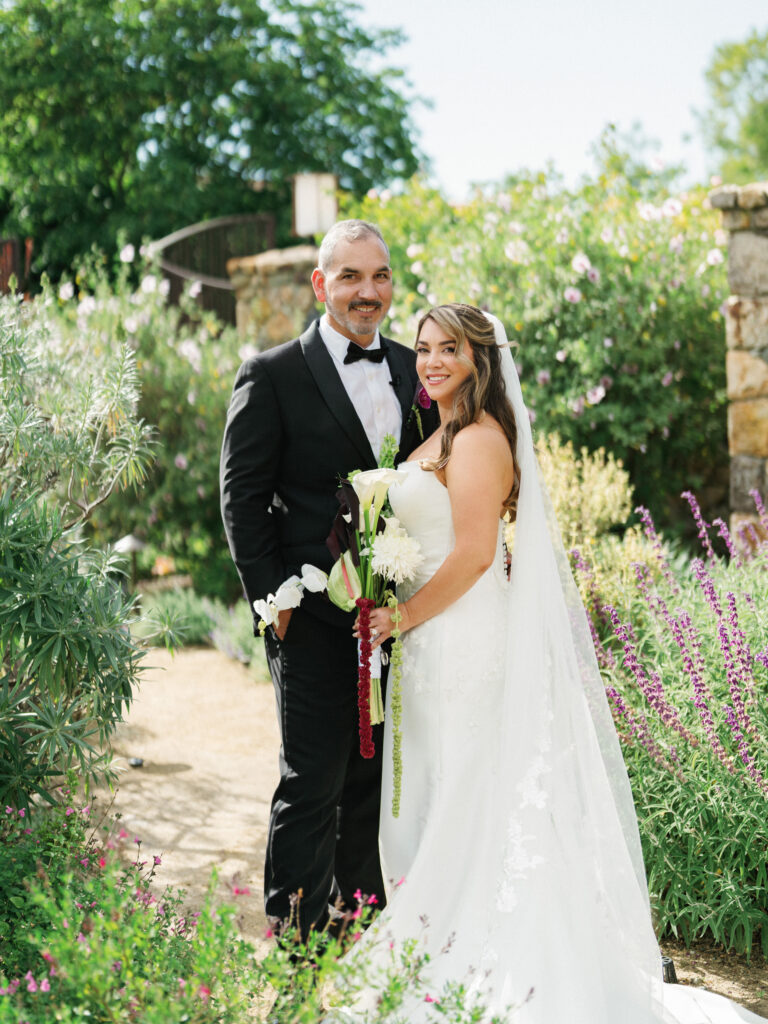 Bride in a fitted lace gown with a long train and groom in a formal black tuxedo standing outdoors surrounded by blooming green bushes.
