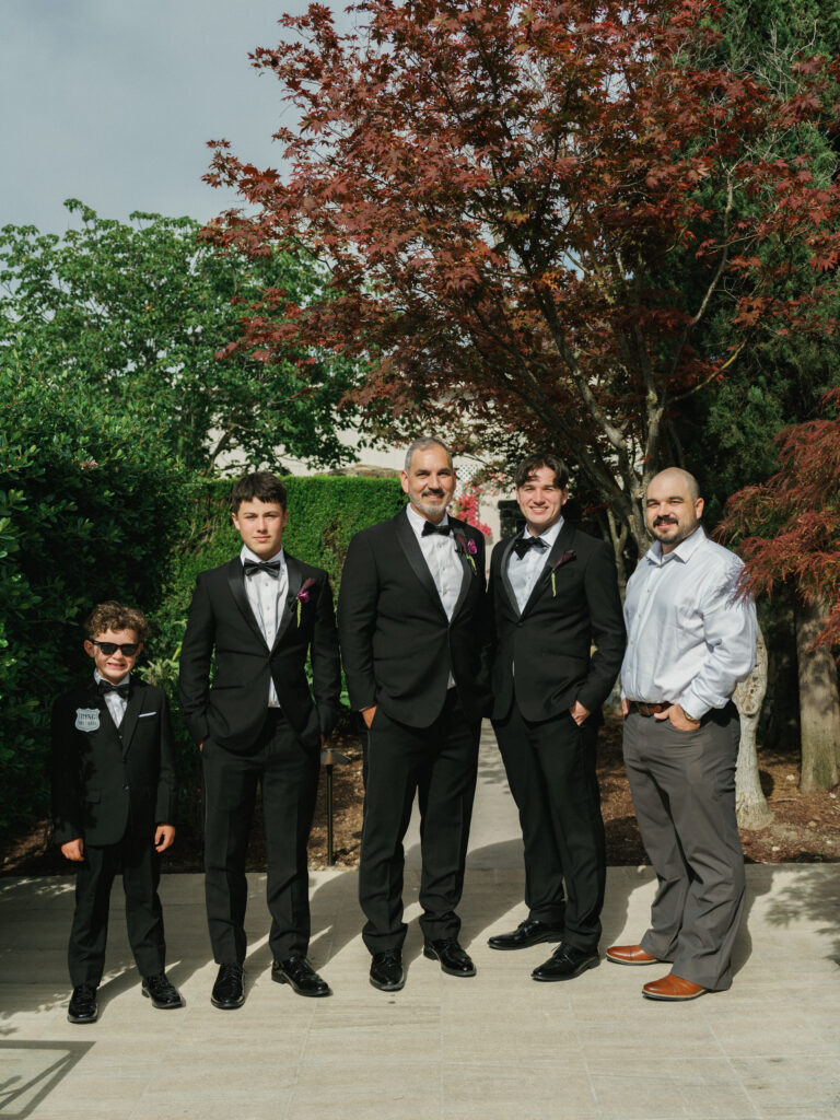 Groom in a black suit with a boutonniere posing with groomsmen in tuxedos and a young ring bearer on a paved garden path outdoors.