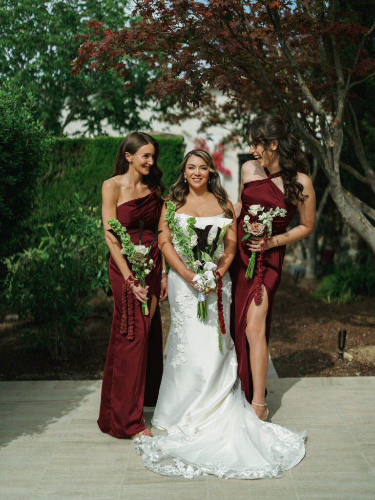 Bride in a fitted lace gown posing with bridesmaids in burgundy satin dresses holding custom calla lily bouquets at a garden wedding.