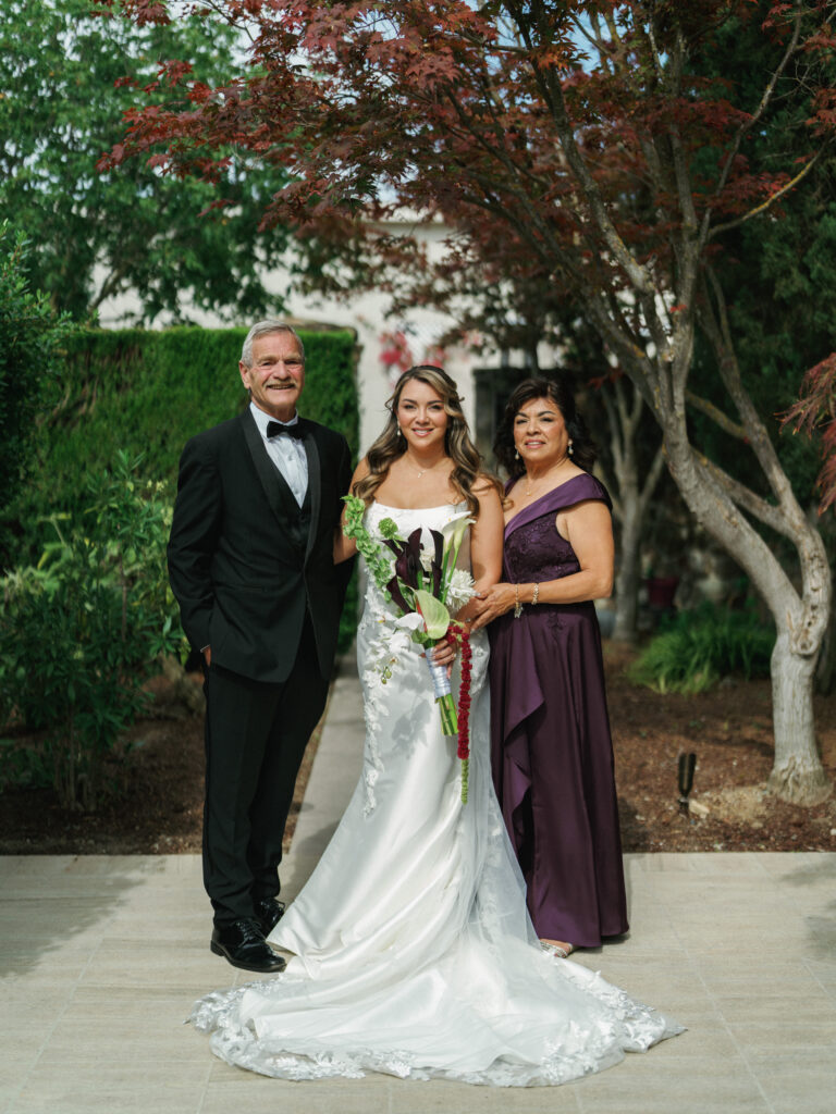 Classic outdoor wedding portrait of a bride with her father in a black tuxedo and mother in a deep purple formal gown at a garden estate reception.