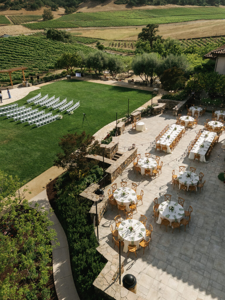 High-angle shot of a vineyard estate wedding reception setup with perfectly arranged dinner tables next to an open ceremony lawn.