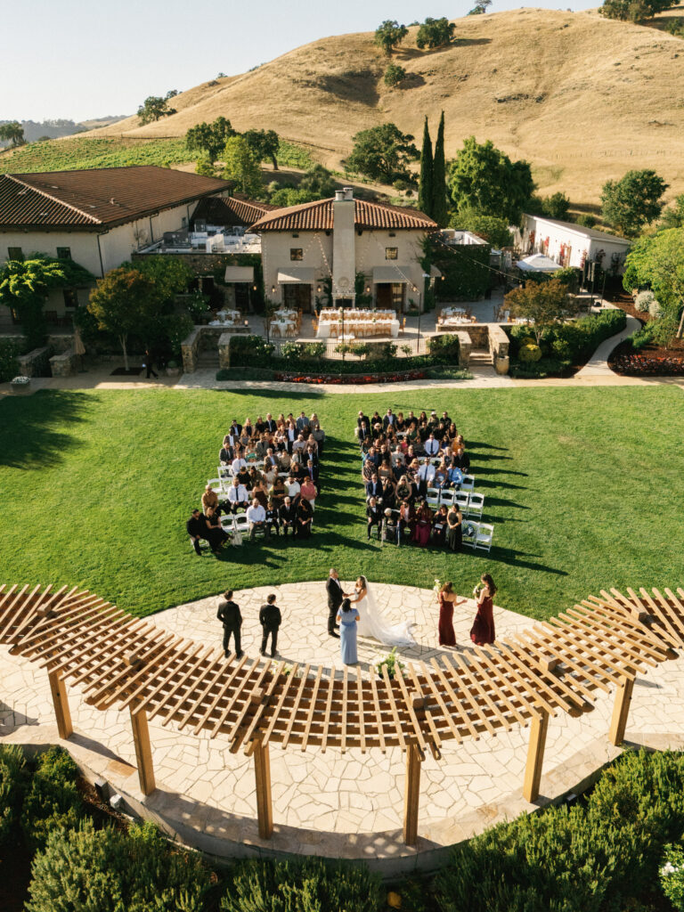 Aerial view of an outdoor wedding ceremony at a luxury California vineyard estate, featuring guests seated on a green lawn by a large wooden pergola.