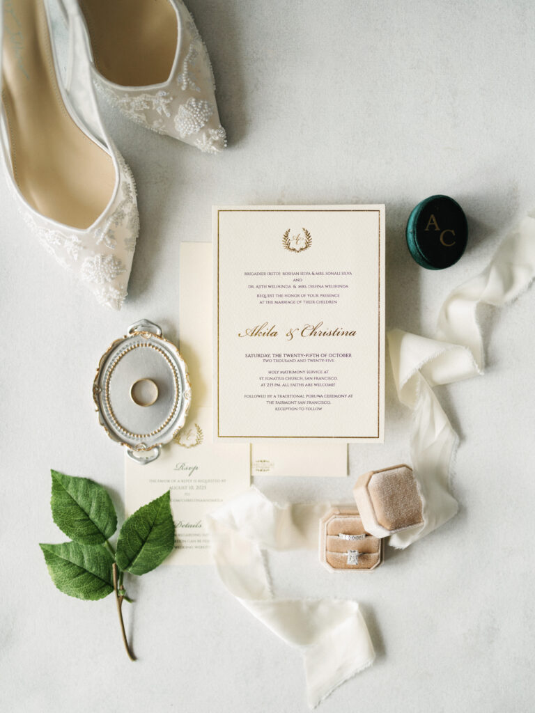 Luxury wedding invitation suite flat lay featuring elegant gold foil calligraphy, lace bridal heels, silk ribbons, and velvet ring boxes on a clean white surface.