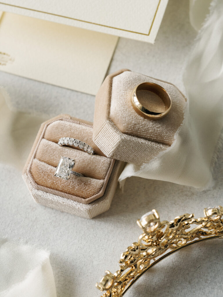 Radiant cut diamond engagement ring and gold band in a beige velvet ring box next to a luxury gold bridal tiara with emerald accents.