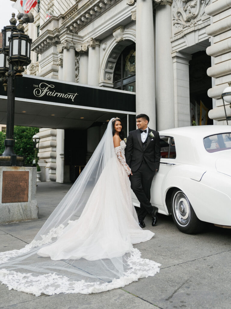 Bride and groom embracing next to a vintage white Rolls-Royce at The Fairmont Hotel wedding entrance in San Francisco, showcasing a lace veil and black tuxedo.
