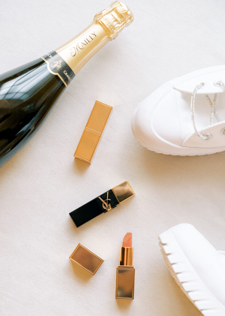 Tom Ford and YSL lipsticks styled with a champagne bottle and white platform bridal sneakers for chic morning wedding detail photos.