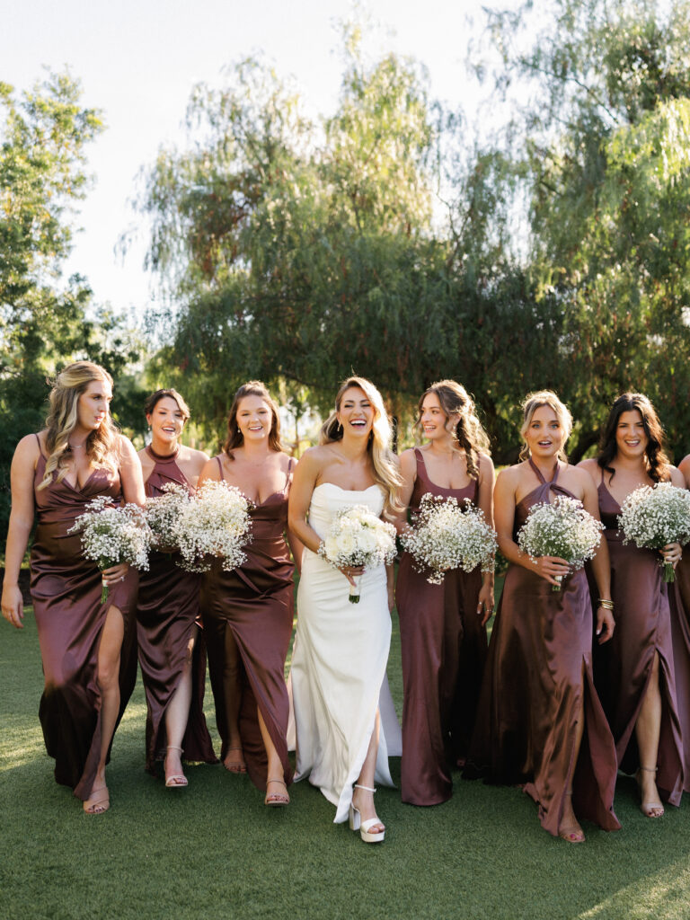 Bride walking on a green lawn with bridesmaids in bronze satin slip dresses holding matching baby's breath bouquets at an outdoor estate wedding.