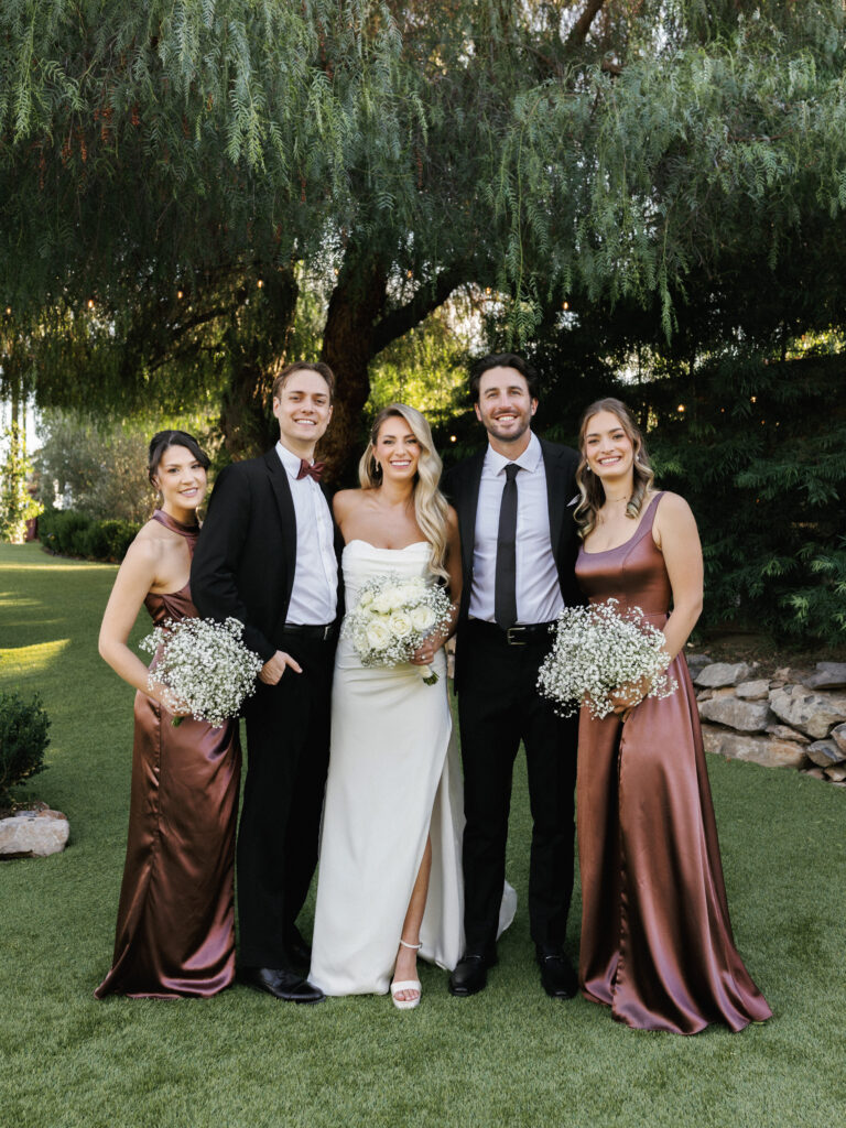 Newlyweds posing with bridesmaids in bronze satin dresses holding baby's breath bouquets on a green lawn at an outdoor garden wedding.