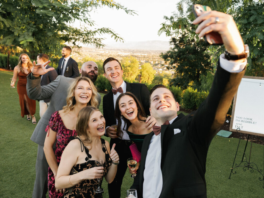 Wedding guests smiling for a phone selfie on a green grass lawn with a valley viewpoint backdrop and a welcome sign on an easel.