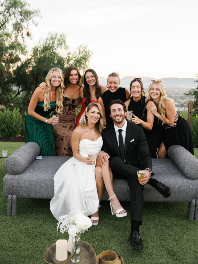 Bride in a strapless dress and groom in a black suit sitting on a modern outdoor sofa, posing with bridesmaids during an estate wedding cocktail hour.