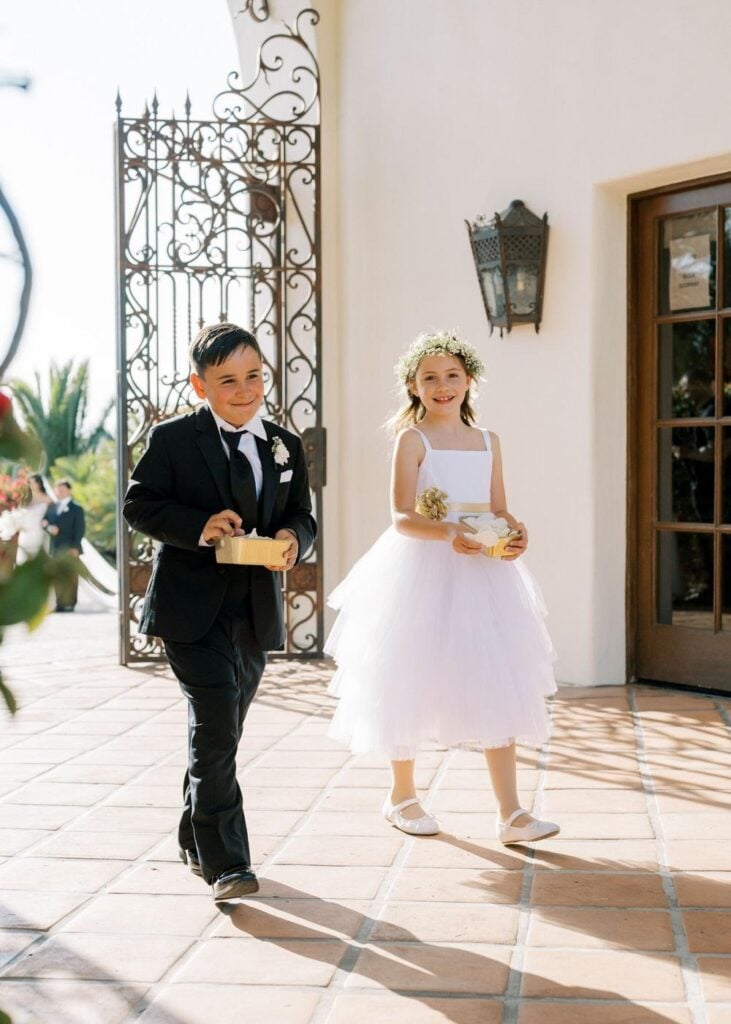 A young ring bearer boy in a black suit and a flower girl in a white tulle dress walking down the aisle during an outdoor ceremony processional.