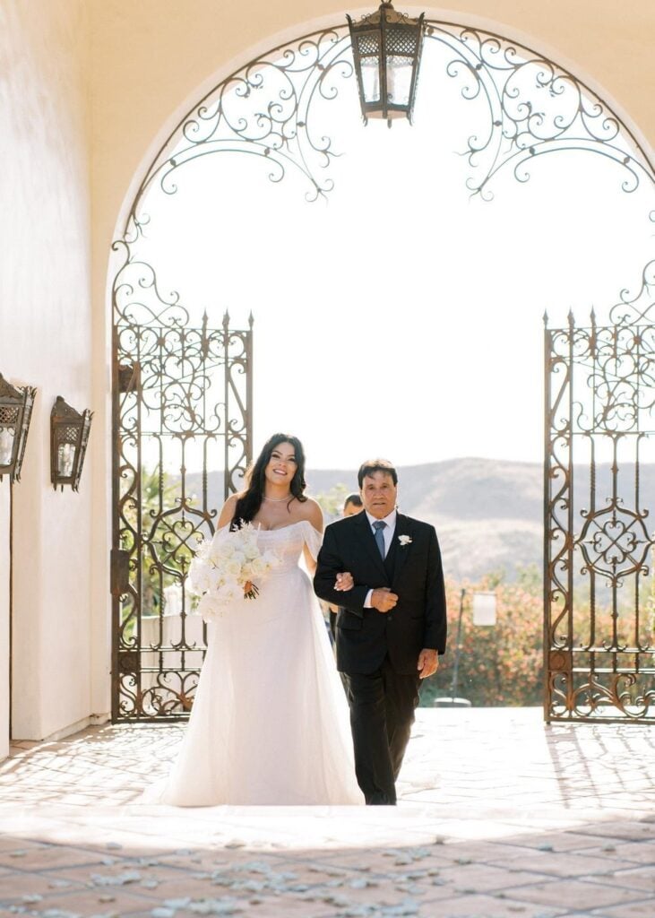 A bride holding a white bouquet and her father walking arm-in-arm through a large arched wrought iron gate for an aisle reveal entrance.
