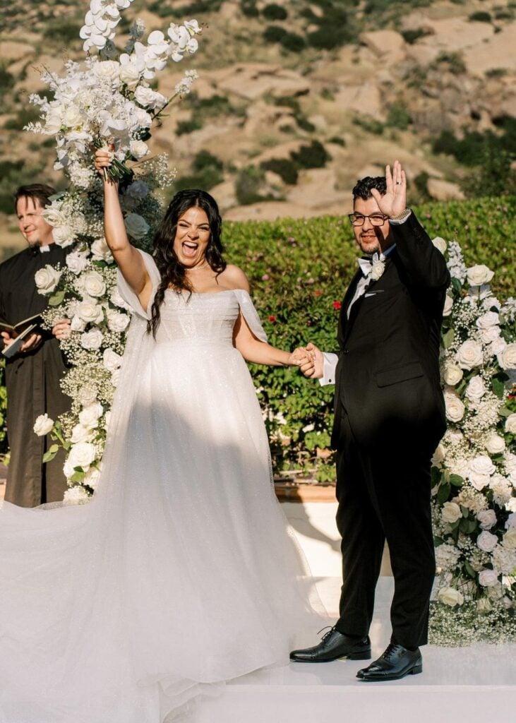 A cheering bride lifting her orchid bouquet and a smiling groom waving his hand while holding hands at a white floral wedding altar.