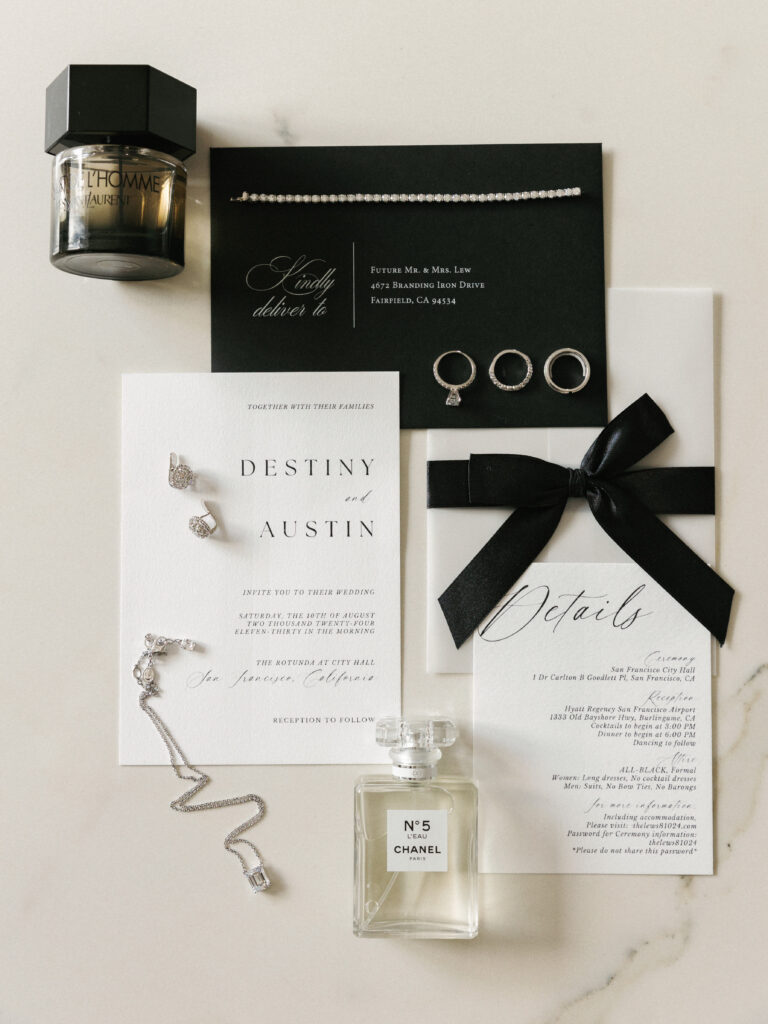Chic black and white wedding invitation suite flat lay with custom calligraphy, a Chanel No 5 perfume bottle, and diamond rings for modern wedding detail photos.