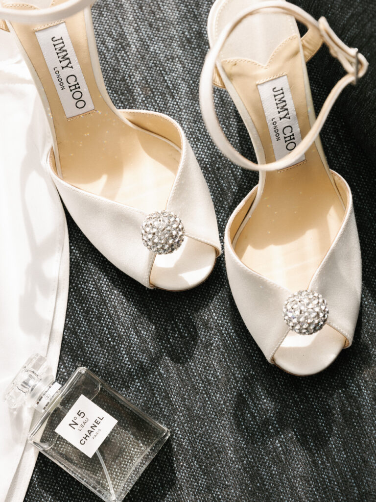 Ivory satin Jimmy Choo bridal sandals with crystal embellishments placed next to a Chanel No 5 perfume bottle on a dark textured surface.