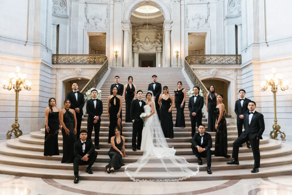 Editorial black tie wedding party portrait on the grand staircase at San Francisco City Hall, featuring a luxury bride in a timeless gown and veil surrounded by bridesmaids in elegant black dresses and groomsmen in classic tuxedos.