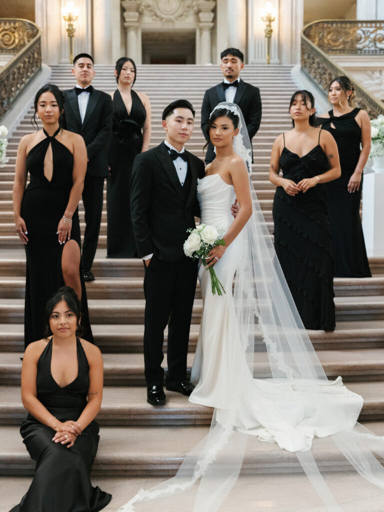 Vogue-inspired bridal party group photo on the grand marble staircase at San Francisco City Hall, showcasing a modern bride in a sleek strapless gown and a long veil with bridesmaids in elegant black dresses.