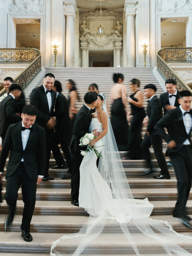 Bride and groom kissing on the Grand Staircase inside San Francisco City Hall with a blurred wedding party
