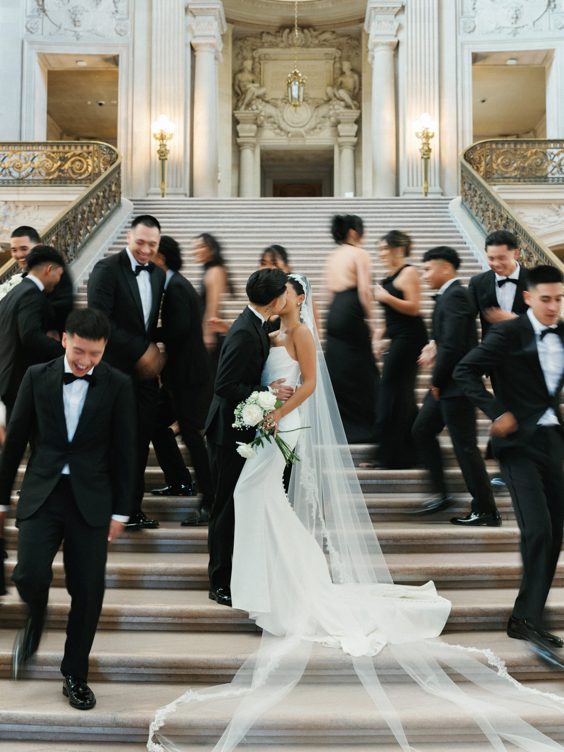 Bride and groom kissing on the Grand Staircase inside San Francisco City Hall with a blurred wedding party
