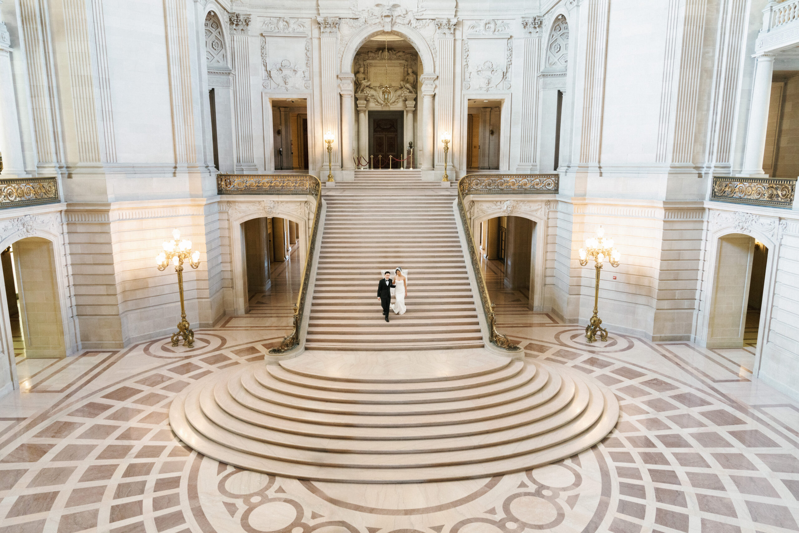 Couple descending a grand staircase in a light-filled hall.