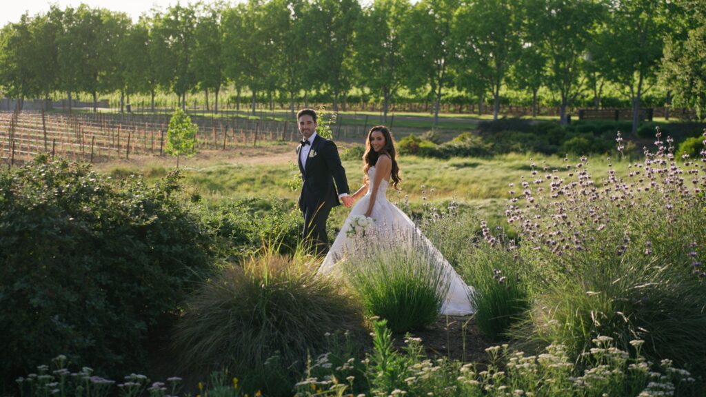 Bride and groom holding hands and walking together through a lush garden path at the Vineyard Area of Greystone Mansion, looking back toward the camera during a golden hour portrait session.