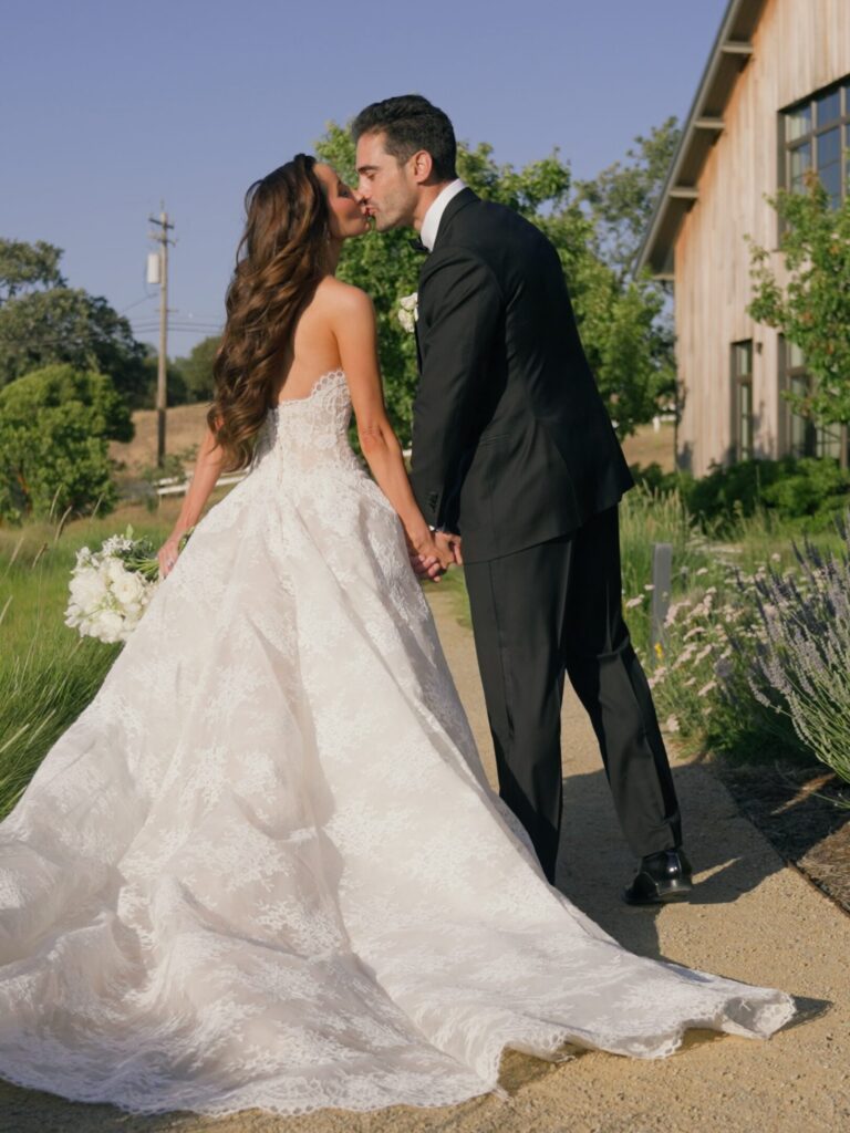 Editorial back view of a bride and groom kissing while standing on a concrete pathway at Sunstone Villa, showcasing the full train of a luxury lace strapless ballgown with lavender bushes nearby.